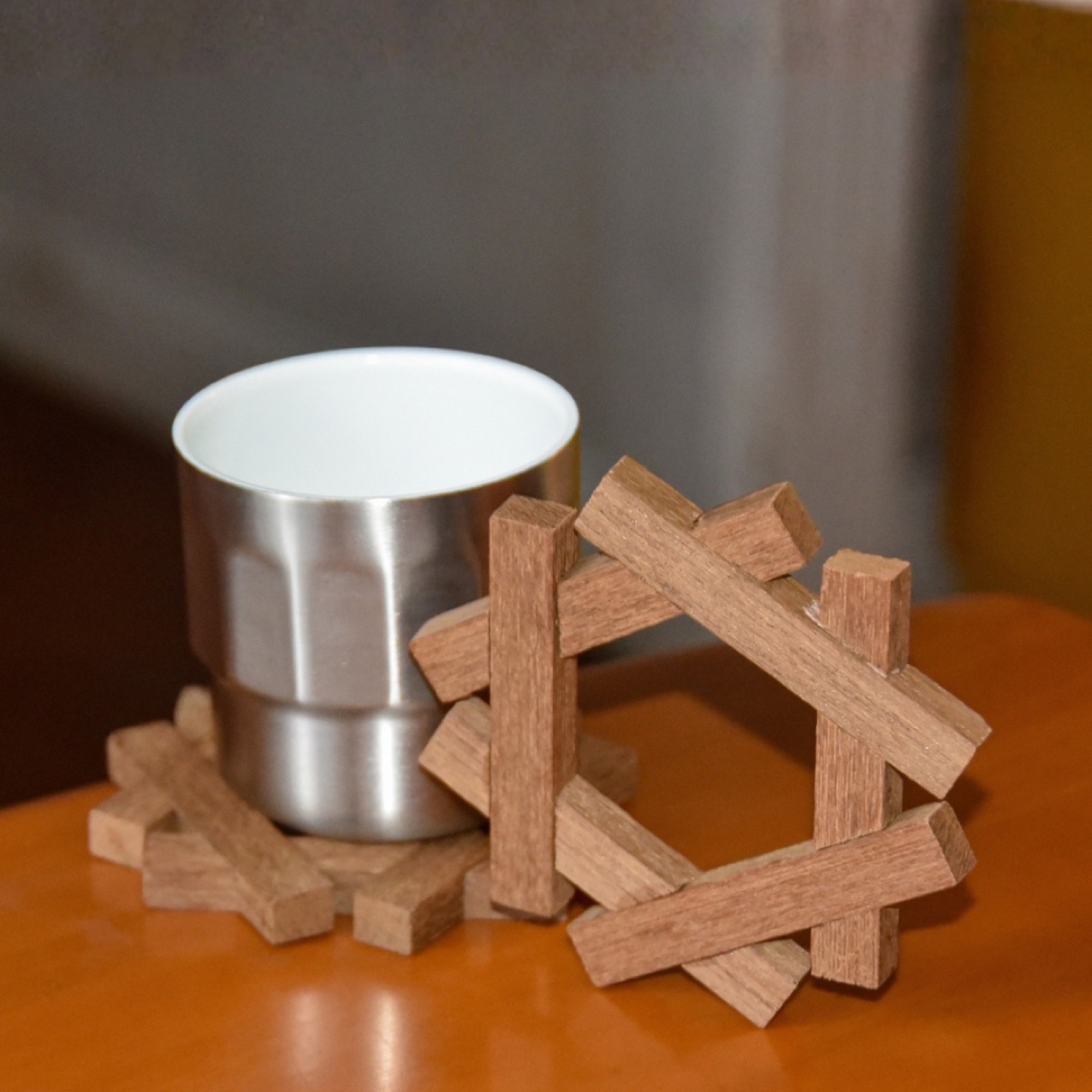 A steel cup resting on an assembled hexagonal wooden coaster, with a second coaster standing upright beside it on a wooden table