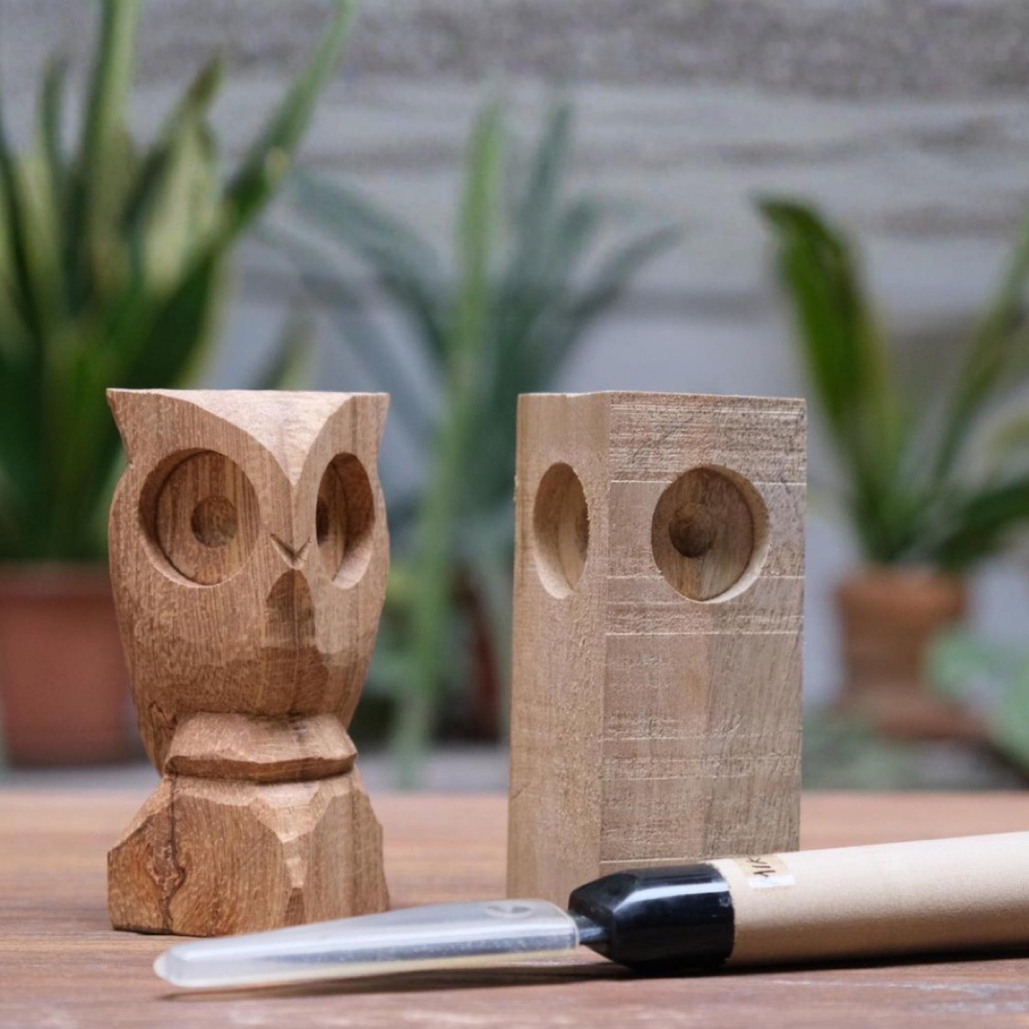 A finished carved wooden owl next to an uncarved timber block and a Japanese whittling knife, on a wooden table with houseplants behind