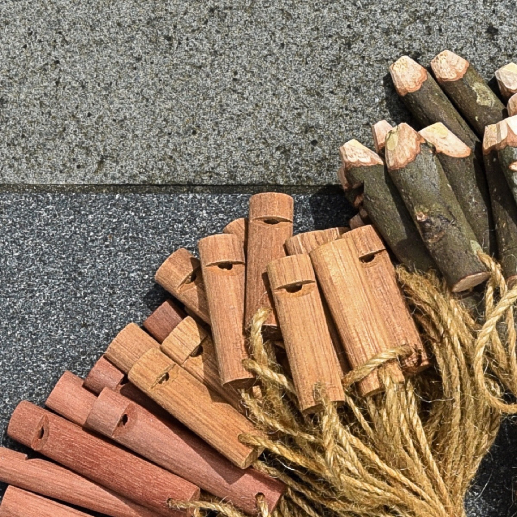 A pile of wooden pocket whistles on a stone surface: reddish minimalist ones at the left, bark-on natural-branch ones at the upper right, all strung with jute cord