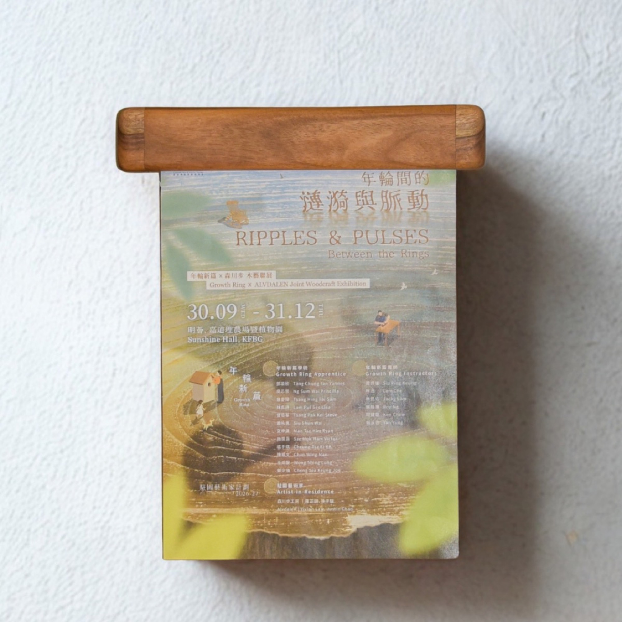The wooden rail mounted on a white wall, gripping a Ripples & Pulses exhibition flyer