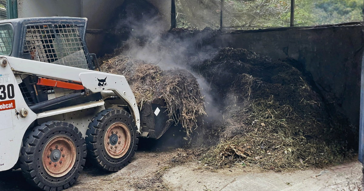 A skid-steer loader turning a steaming compost windrow inside the open-sided compost shed